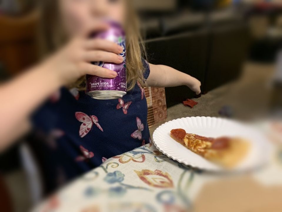 Little girl sitting at the dining room table enjoying a slice of pizza and a soda.